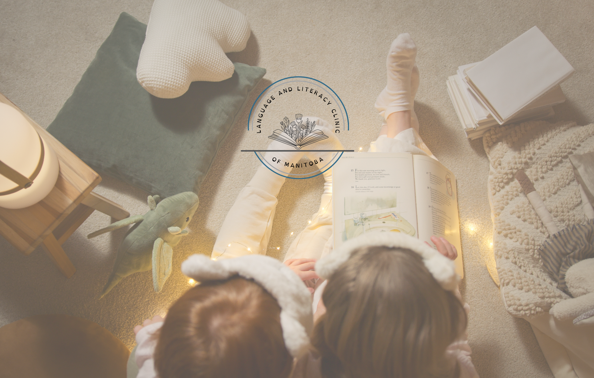 back of 2 children's heads sitting on a floor while reading a book, with twinkle lights wrapped around them. The Language and Literacy Clinic of Manitoba logo can be seen at the top of the image. 